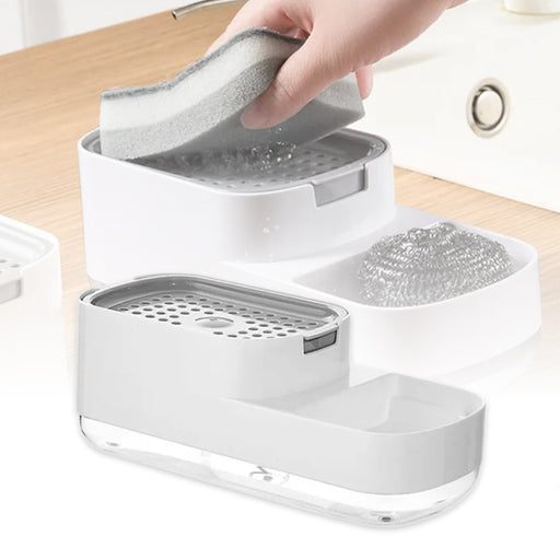 A white kitchen sink organizer with a soap dispenser, featuring a sponge holder and an additional storage tray, placed on a wooden surface.