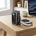 A portable power bank by Acefast sits on a wooden desk, connected to a smartphone via a black cable. The scene is a styled workspace with a laptop, a white coffee mug, and a small potted succulent in the background.