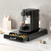 Black coffee machine with a drawer full of capsules on a kitchen counter.