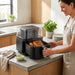 A smiling woman in an apron opens the drawer of a black air fryer on a concrete countertop to reveal a whole roasted, golden-brown chicken.