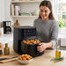 A woman in a grey sweater smiles as she pulls open the drawer of a black GreenPan air fryer, revealing a basket full of cooked chicken pieces. 