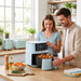A smiling couple using a Smokey Sky blue GreenPan air fryer together in a bright modern kitchen, with one person operating the touchscreen and the other holding open the drawer filled with cooked food.