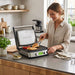 A smiling woman uses tongs to flip grilled chicken breasts on an open, multi-purpose countertop grill from GreenPan. 