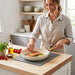 A smiling woman wears an apron while arranging thyme sprigs over seared salmon with lemons in a cream frying pan on an induction cooktop.