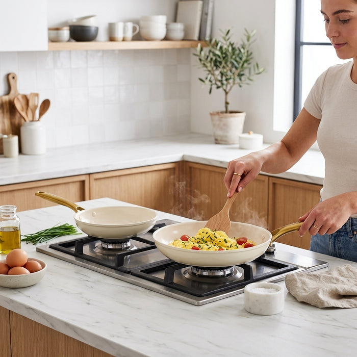 Person cooking scrambled eggs with tomatoes in a cream skillet with a gold handle on a kitchen stovetop.
