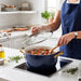 A person stirring a simmering stew in a navy blue GreenPan casserole pot with gold handles on a modern stovetop.