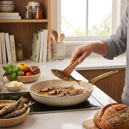 A hand uses wooden tongs to turn a whole fried fish in fry pan.