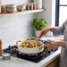 A person in a linen shirt uses a gold-colored spatula to cook chicken and vegetable stir-fry in a white sauté pan on a gas stovetop in a modern kitchen.