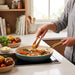 A person uses wooden tongs to stir tofu and vegetables in a smokey blue pan.