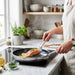 A person in a beige linen apron flips a seared salmon fillet in a stainless steel frypan on an induction cooktop.