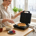 A woman smiles as she uses a spatula to lift a golden Belgian waffle from an open, black four-quadrant waffle maker on a kitchen counter.