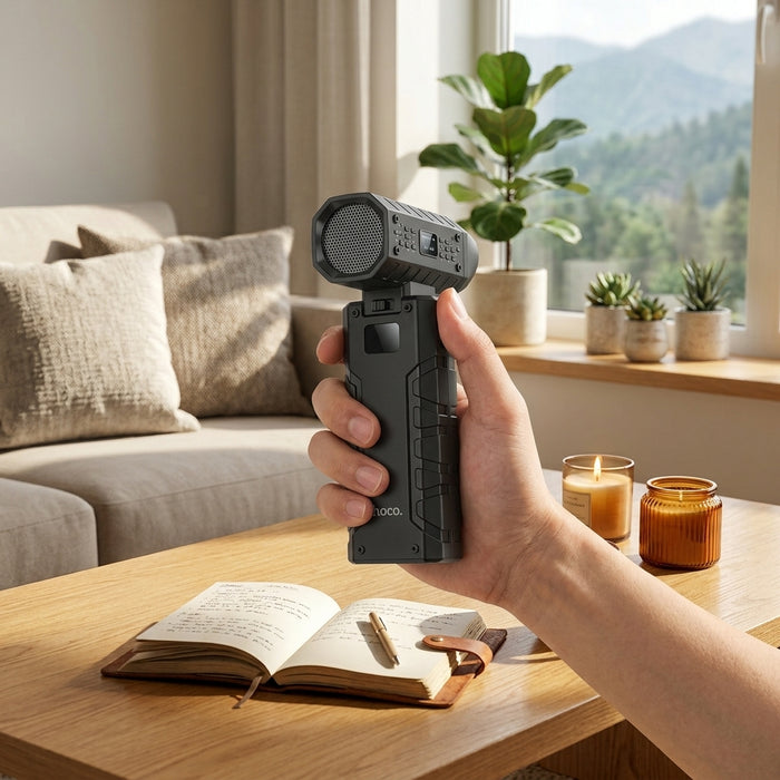 A hand holding a black portable rechargeable fan over a wooden coffee table next to an open journal and a lit candle.