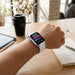 A person's wrist wearing a  smartwatch with a detailed data screen, resting on a wooden table alongside a tablet, coffee cup, keys, and notebook in a bright room.