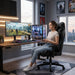 A young woman sits in gaming chair, holding a controller and looking at an ultrawide monitor displaying an open-world fantasy game in a dedicated gaming room.