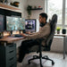 A young man sitting in a grey Playmax gaming chair at a modern, organized desk setup, focused on playing a video game on a large monitor.