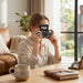 A young woman holds a retro-style digital camera to her face, sitting at a sunlit wooden table. 