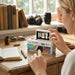 A woman sitting at a wooden desk adjusting the flip-up screen of a white retro mini digital camera.