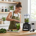 A woman prepares a green smoothie using a Vitamix Ascent A3500i blender in a bright, modern kitchen.
