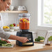 A woman uses a Matt Blue Vitamix blender to prepare a fruit smoothie in a bright, modern kitchen.