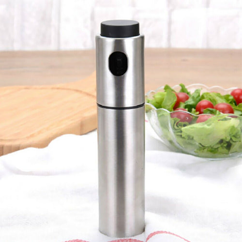 
A close-up product photo of a stainless steel oil spray bottle with a black cap and nozzle button, placed on a white cloth. A wooden cutting board and a glass bowl of fresh salad with lettuce and cherry tomatoes are blurred in the background, suggesting use for kitchen oil misting or cooking spray.
