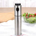 
A close-up product photo of a stainless steel oil spray bottle with a black cap and nozzle button, placed on a white cloth. A wooden cutting board and a glass bowl of fresh salad with lettuce and cherry tomatoes are blurred in the background, suggesting use for kitchen oil misting or cooking spray.