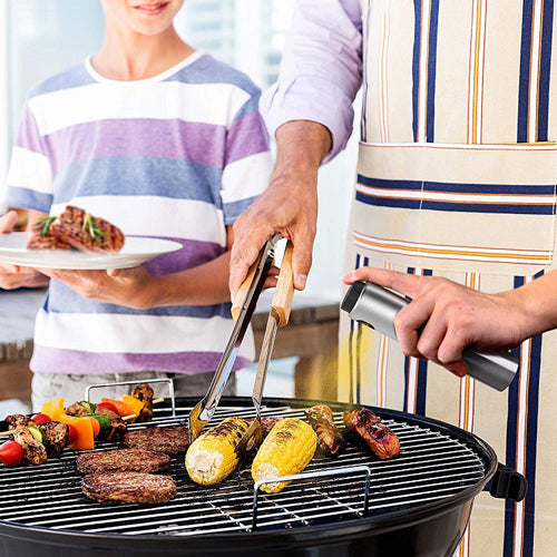 
A father and son are seen grilling outdoors. The man, wearing an apron, is using a pair of metal grilling tongs with wooden handles to turn food (burgers and corn cobs) on a round charcoal grill, while also holding a stainless steel oil spray bottle in his other hand.