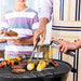 
A father and son are seen grilling outdoors. The man, wearing an apron, is using a pair of metal grilling tongs with wooden handles to turn food (burgers and corn cobs) on a round charcoal grill, while also holding a stainless steel oil spray bottle in his other hand.