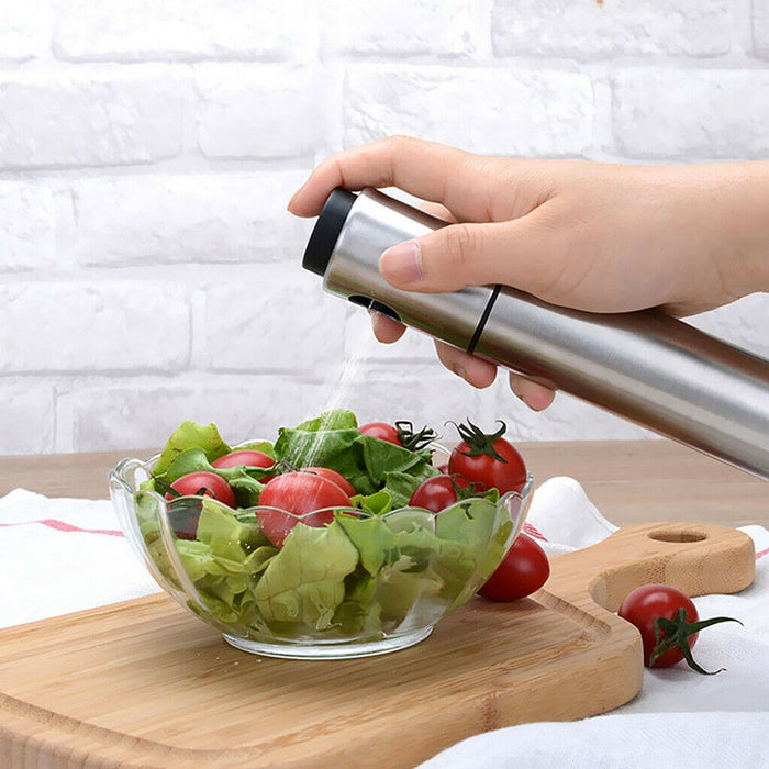 
A close-up shot of a hand using a stainless steel oil spray bottle to mist dressing onto a clear glass bowl of fresh salad, which contains crisp lettuce and cherry tomatoes. The bowl rests on a wooden cutting board with a white brick wall background, highlighting the cooking oil mister in use.