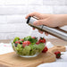 
A close-up shot of a hand using a stainless steel oil spray bottle to mist dressing onto a clear glass bowl of fresh salad, which contains crisp lettuce and cherry tomatoes. The bowl rests on a wooden cutting board with a white brick wall background, highlighting the cooking oil mister in use.