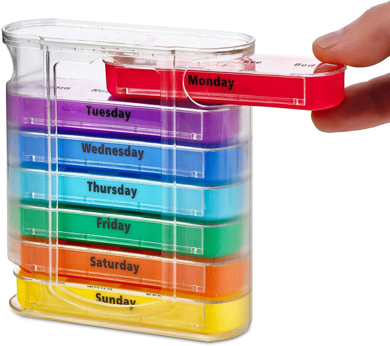 A clear plastic, vertical pill organizer holding seven colorful, stacked daily pill trays (Monday through Sunday). A hand is pulling out the red 'Monday' tray from the top of the container. Each tray is a different color of the rainbow and labeled with the day of the week in black text.