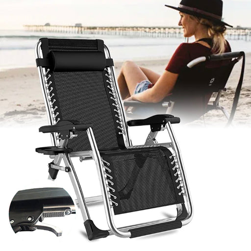 A black deluxe folding beach lounger with adjustable headrest and footrest, displayed in a reclined position, with an additional cushion for back support. The lounger is shown both in a folded and an unfolded position, and is placed in an outdoor setting with a person seated in the background.