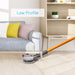 A product demonstration image showing a gray and clear motorized mop head attachment connected to an orange stick vacuum pole, cleaning the light wooden floor underneath a light gray sofa. Text overlay reads: "Low Profile," highlighting the mop head's slim design for cleaning hard-to-reach areas.
