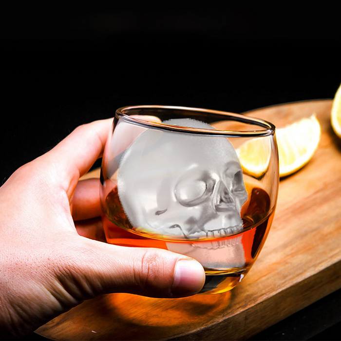 Hand holding a glass with a skull design filled with a amber liquid on a wooden surface.