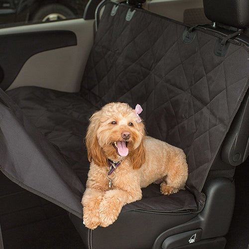 
A photo showing a small, happy, light-brown dog (likely a Cockapoo or similar breed) with a pink bow sitting on a black, quilted, waterproof pet hammock/car seat cover installed on the rear bench seat of a vehicle. The cover protects the seat and backrest.