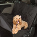 
A photo showing a small, happy, light-brown dog (likely a Cockapoo or similar breed) with a pink bow sitting on a black, quilted, waterproof pet hammock/car seat cover installed on the rear bench seat of a vehicle. The cover protects the seat and backrest.