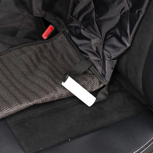A close-up of a white, cylindrical seat anchor (or seat wedge) that is part of a black car seat cover or pet hammock. The anchor is inserted into the crease between the backrest and the seat cushion. The cover is made of black, quilted fabric and a black mesh material near the anchor. A red seat belt buckle is partially visible in the upper left corner.