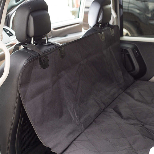 A close-up photo of a black, quilted, waterproof pet hammock/car seat cover installed on the rear bench seat of a vehicle. The cover is secured around the headrests with adjustable straps and protects the backrest and bench seat.