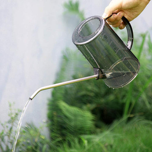 A clear plastic watering can with a stainless steel spout being held by a person, water is flowing from the spout into the soil.