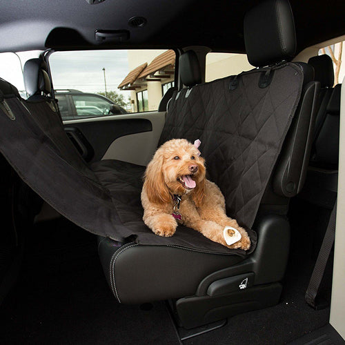 A small, happy-looking, light brown or apricot-colored Doodle-type dog (such as a Cockapoo or Goldendoodle) is sitting on a black, quilted car seat cover. The cover is installed in the second row of a vehicle, appearing to be a pet hammock setup, covering the seat and looping up to the back of the front seats .