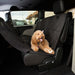 A small, happy-looking, light brown or apricot-colored Doodle-type dog (such as a Cockapoo or Goldendoodle) is sitting on a black, quilted car seat cover. The cover is installed in the second row of a vehicle, appearing to be a pet hammock setup, covering the seat and looping up to the back of the front seats .