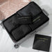An open silver rolling suitcase is organized using black travel packing cubes and laundry pouches, all labeled with gold text. The cubes hold dark clothing, demonstrating a compact travel organization system on a white quilted surface.