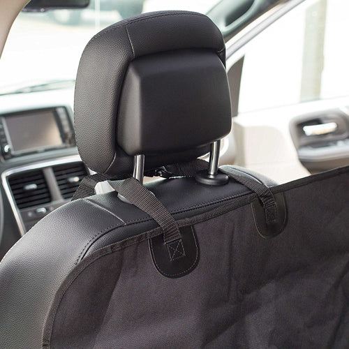 A close-up view of a black fabric car seat protector or pet hammock secured to the back of a black leather car seat. Black adjustable straps loop around the metal posts and base of the headrest to hold the cover in place. The car's interior, including the dashboard and door panel, is visible in the background.