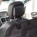 A close-up view of a black fabric car seat protector or pet hammock secured to the back of a black leather car seat. Black adjustable straps loop around the metal posts and base of the headrest to hold the cover in place. The car's interior, including the dashboard and door panel, is visible in the background.
