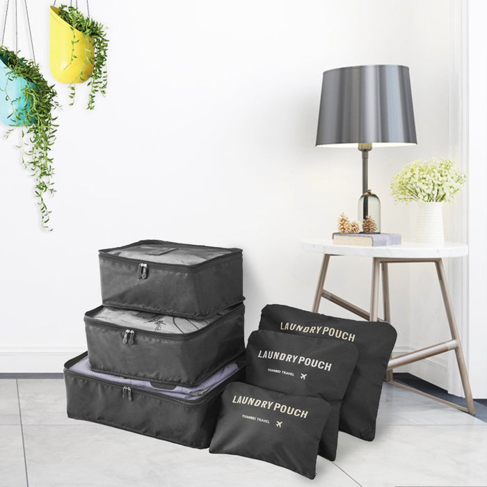 A staged lifestyle photo showing a set of black travel packing cubes and laundry pouches stacked on the floor in a modern room. The three mesh organizer bags and three flat laundry pouches are positioned next to a small white side table with a gray lamp and white flowers. Hanging plants in vibrant yellow and blue pots decorate the white wall in the upper left corner.
