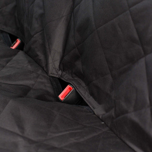 A close-up of a black, quilted fabric car seat cover showing an opening for a seat belt buckle. A red and black seat belt buckle is visible poking through a slit in the fabric. A second, partially visible buckle is to the left, indicating the cover is designed to allow seat belts to be used.